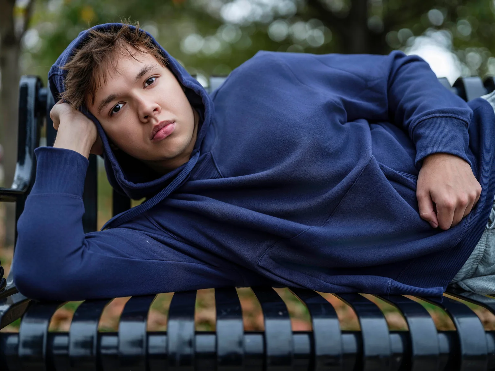 Max Cooper III in a blue hoodie lying on a park bench, looking into the camera